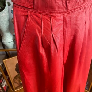 May include: Close-up of red leather pants with a high waist and pleated front. The pants have a wide belt and pockets. The leather appears smooth and the color is a vibrant red.