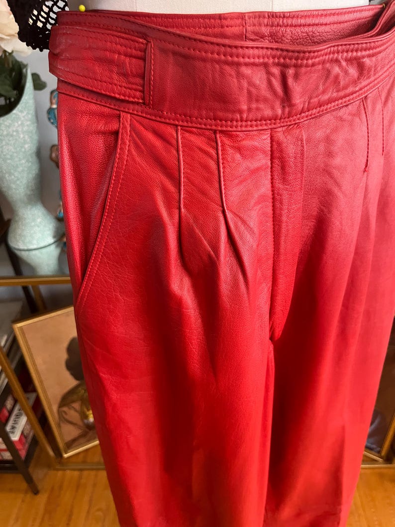 May include: Close-up of red leather pants with a high waist and pleated front. The pants have a wide belt and pockets. The leather appears smooth and the color is a vibrant red.