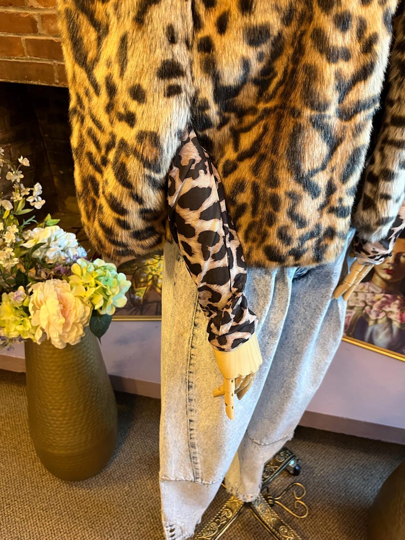 K&ouml;nnte beinhalten: Eine kurz geschnittene Jacke aus Kunstfell mit braun-schwarzem Leopardenmuster, kombiniert mit einem lang&auml;rmeligen Oberteil im passenden Leopardenmuster und einer hellblauen Jeans aus Denim, pr&auml;sentiert auf einer Schaufensterpuppe.
