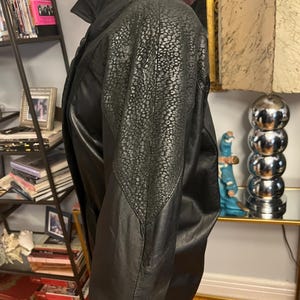 May include: Black leather jacket with a textured shoulder panel. The jacket features a high collar and a classic design. The leather appears to be in good condition, suggesting a vintage or well-maintained piece.