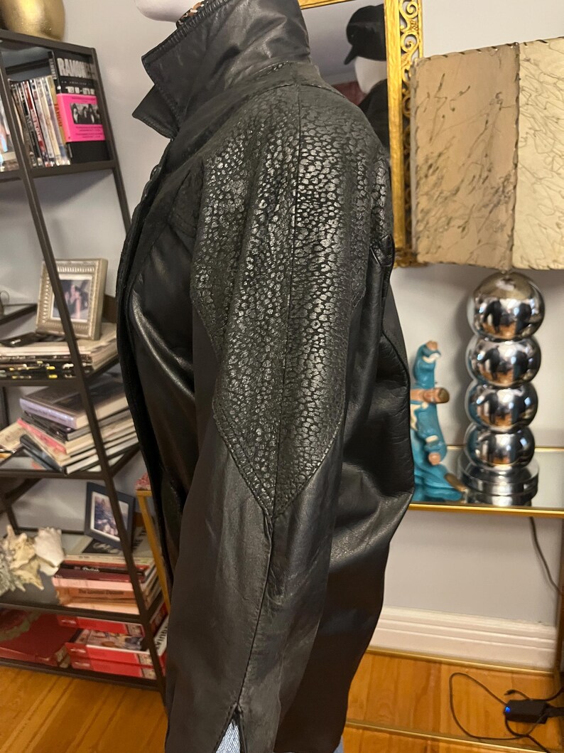 May include: Black leather jacket with a textured shoulder panel. The jacket features a high collar and a classic design. The leather appears to be in good condition, suggesting a vintage or well-maintained piece.