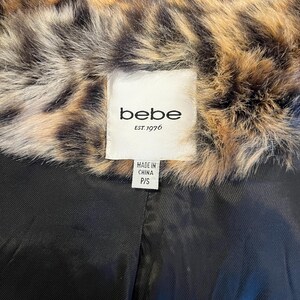 K&ouml;nnte beinhalten: Ein Kleidungsst&uuml;ck mit einem braun-schwarzen Kunstfellkragen im Leopardenmuster und einem einfarbig schwarzen Innenfutter. Ein wei&szlig;es Stoffetikett am Kragen zeigt den Text "bebe EST. 1976" sowie ein kleines Etikett mit "MADE IN CHINA P/S".