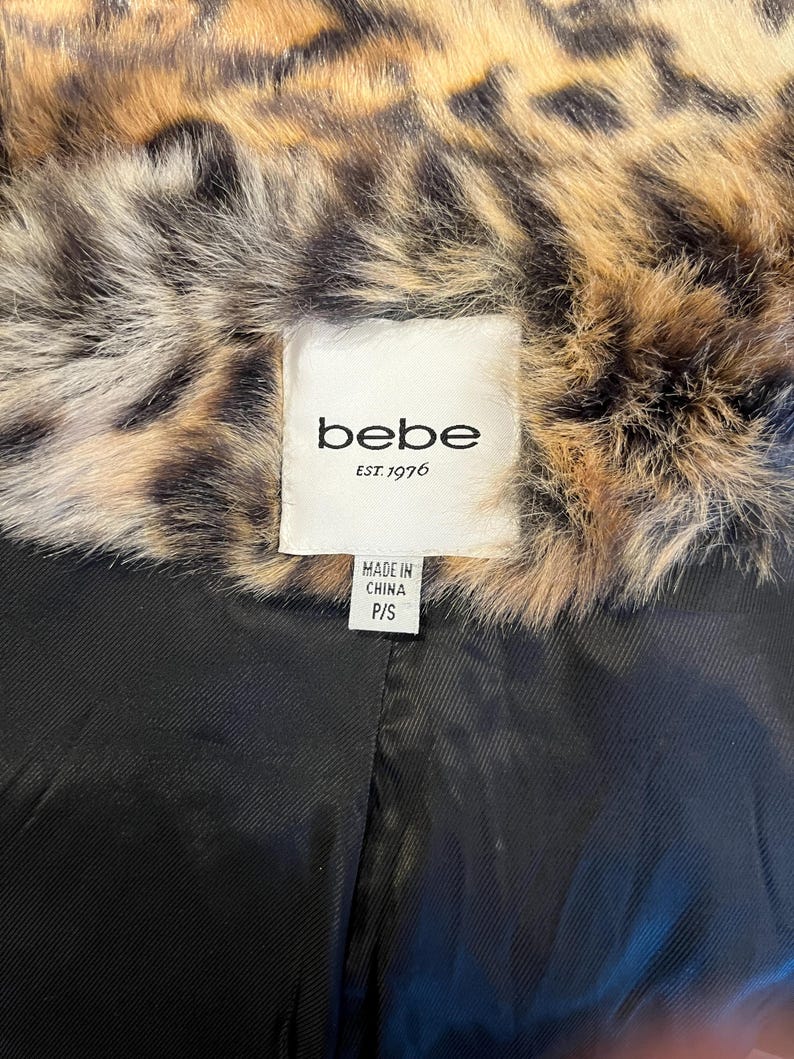 K&ouml;nnte beinhalten: Ein Kleidungsst&uuml;ck mit einem braun-schwarzen Kunstfellkragen im Leopardenmuster und einem einfarbig schwarzen Innenfutter. Ein wei&szlig;es Stoffetikett am Kragen zeigt den Text "bebe EST. 1976" sowie ein kleines Etikett mit "MADE IN CHINA P/S".