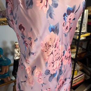 May include: A sleeveless, lilac-colored dress with a floral pattern. The dress features large pink roses with blue and grey leaves. The fabric has a silky sheen, and the dress is displayed on a mannequin.
