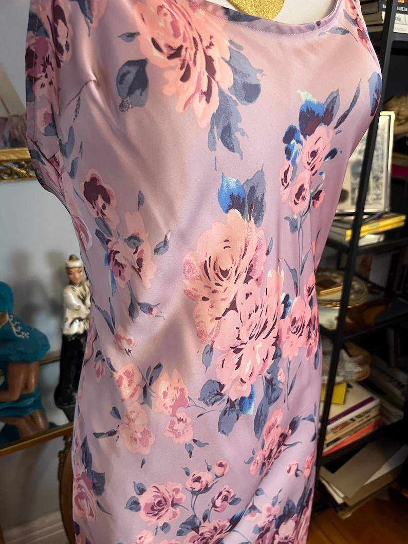 May include: A sleeveless, lilac-colored dress with a floral pattern. The dress features large pink roses with blue and grey leaves. The fabric has a silky sheen, and the dress is displayed on a mannequin.