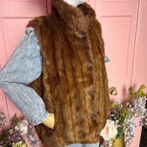 May include: A brown fur vest with a high collar and three dark buttons. The vest is worn over a light blue denim jacket. The mannequin is wearing a light-colored hat. The background includes a pink door and a gold vase with flowers.