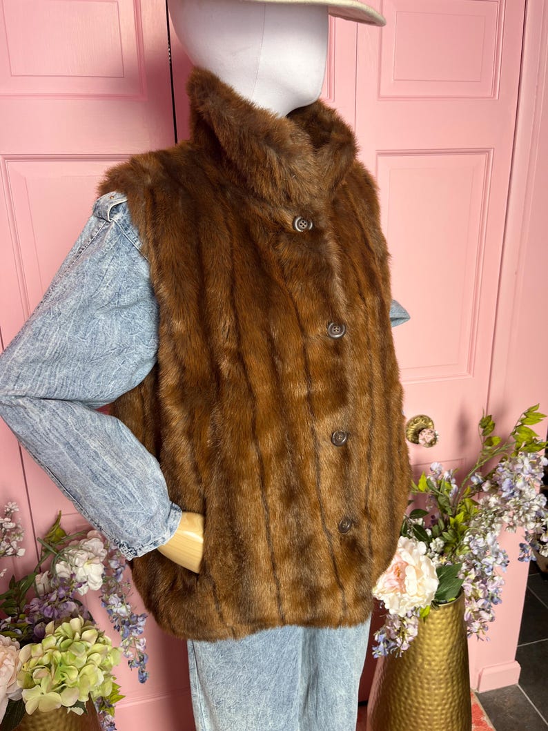 May include: A brown fur vest with a high collar and three dark buttons. The vest is worn over a light blue denim jacket. The mannequin is wearing a light-colored hat. The background includes a pink door and a gold vase with flowers.
