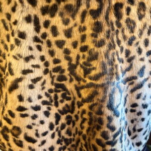 K&ouml;nnte beinhalten: Ein Kleidungsst&uuml;ck mit einer dichten Kunstfelltextur mit kurzem Flor und einem sich wiederholenden Leopardenmuster. Das Design besteht aus unregelm&auml;&szlig;igen schwarzen und dunkelbraunen Flecken auf einem hellbraunen und cremefarbenen Hintergrund, was eine naturalistisch von Tieren inspirierte &Auml;sthetik auf der gesamten sichtbaren Stoffoberfl&auml;che erzeugt.