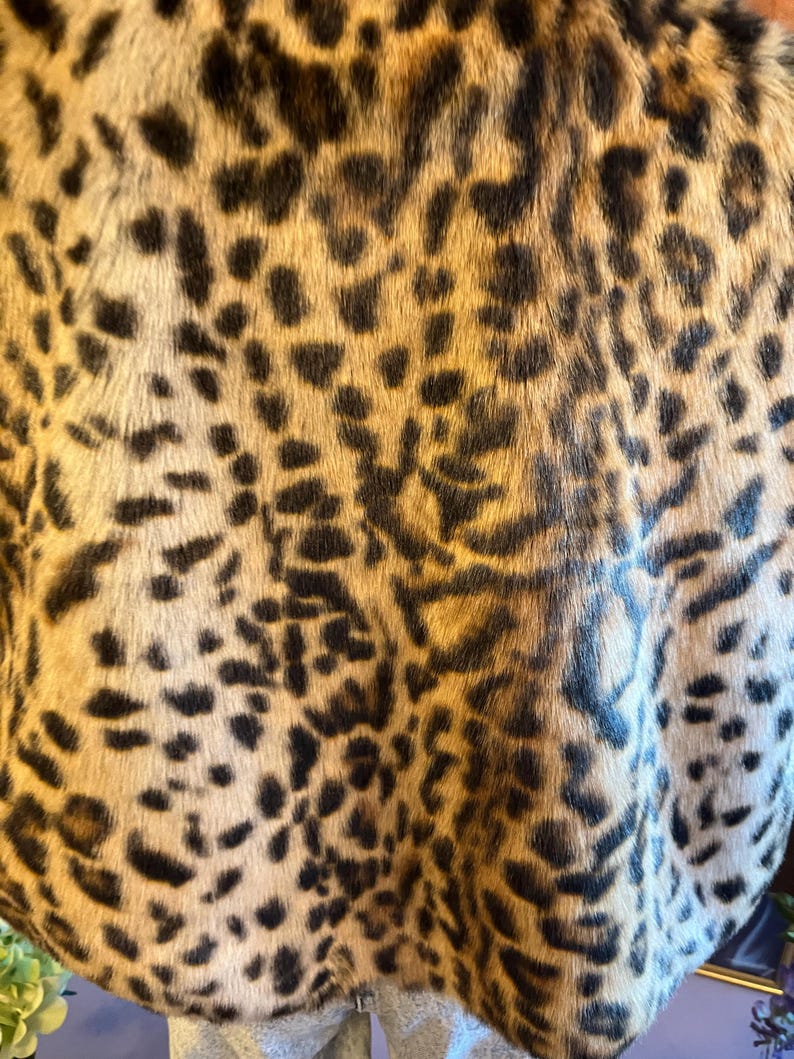 K&ouml;nnte beinhalten: Ein Kleidungsst&uuml;ck mit einer dichten Kunstfelltextur mit kurzem Flor und einem sich wiederholenden Leopardenmuster. Das Design besteht aus unregelm&auml;&szlig;igen schwarzen und dunkelbraunen Flecken auf einem hellbraunen und cremefarbenen Hintergrund, was eine naturalistisch von Tieren inspirierte &Auml;sthetik auf der gesamten sichtbaren Stoffoberfl&auml;che erzeugt.