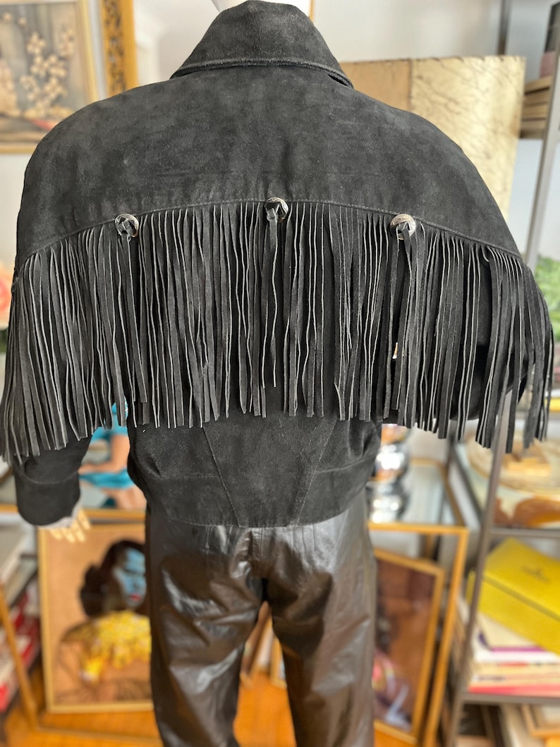May include: Black suede jacket with fringe detail across the back. The jacket has a classic collar and silver-toned button accents. The fringe is long and hangs down from the shoulders. A stylish piece of outerwear.