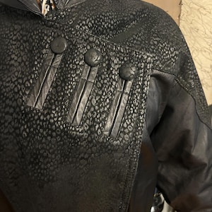 May include: Black leather jacket with a unique leopard print pattern on the shoulders and upper chest. The jacket features three large, round buttons and decorative vertical leather strips. The collar is a classic design, and the jacket appears to be vintage.