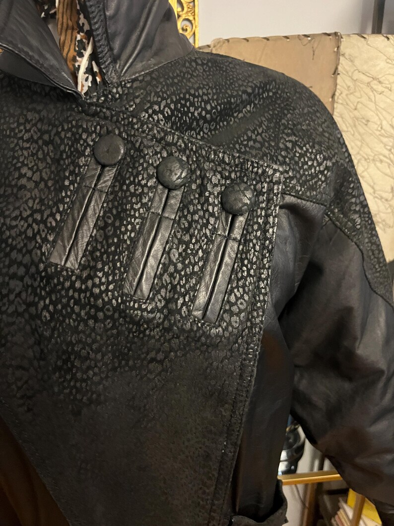 May include: Black leather jacket with a unique leopard print pattern on the shoulders and upper chest. The jacket features three large, round buttons and decorative vertical leather strips. The collar is a classic design, and the jacket appears to be vintage.