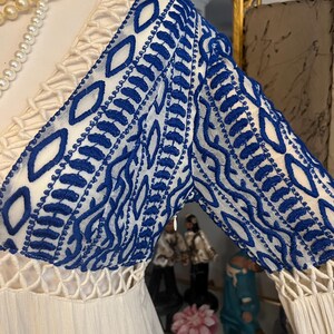 Pu&ograve; includere: Abito color crema con maniche ricamate blu. Le maniche presentano un motivo dettagliato di cuciture blu su sfondo bianco. L'abito ha uno scollo a V e un corpetto strutturato.