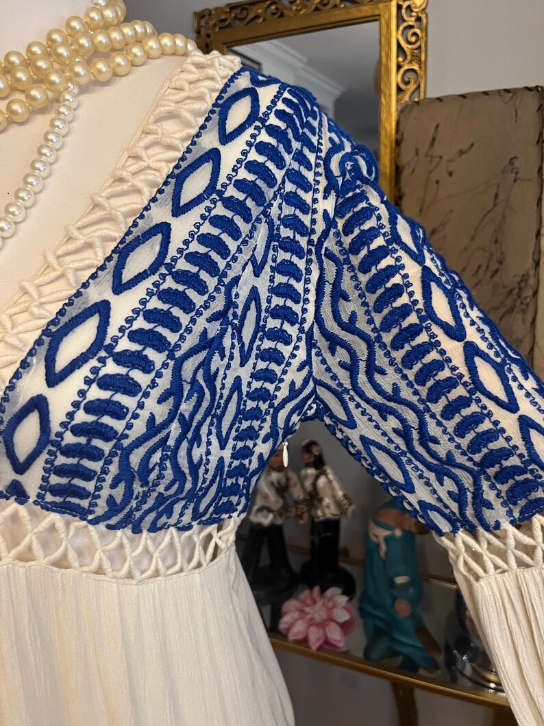 Pu&ograve; includere: Abito color crema con maniche ricamate blu. Le maniche presentano un motivo dettagliato di cuciture blu su sfondo bianco. L'abito ha uno scollo a V e un corpetto strutturato.