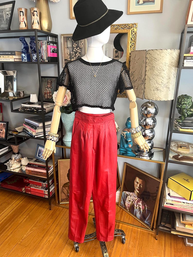 May include: A mannequin displays a black mesh crop top and high-waisted red pants. The outfit is accessorized with a black hat with a gold band and bracelets. The pants are pleated and have a wide leg. The mannequin is on a rolling stand.
