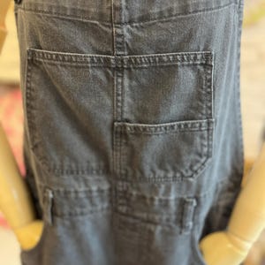 May include: Close-up of a pair of charcoal gray denim overalls. The overalls feature multiple pockets, including a large back pocket and smaller front pockets. The straps are secured with dark metal buttons. The overall design is casual and practical.
