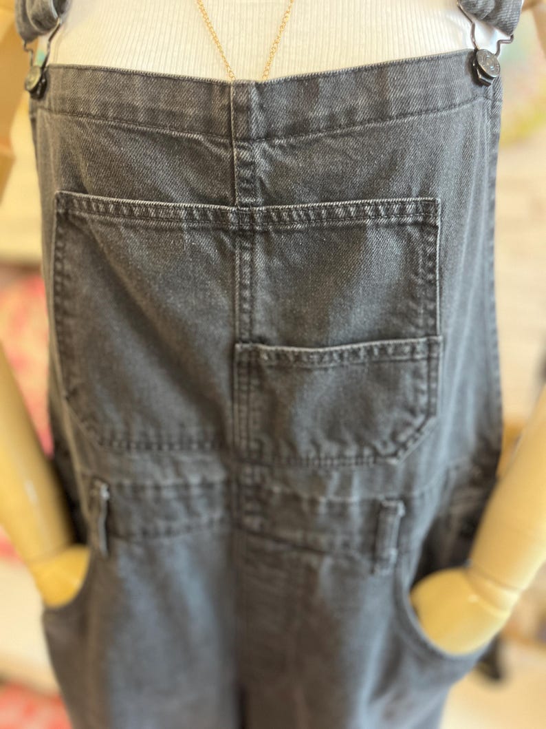 May include: Close-up of a pair of charcoal gray denim overalls. The overalls feature multiple pockets, including a large back pocket and smaller front pockets. The straps are secured with dark metal buttons. The overall design is casual and practical.