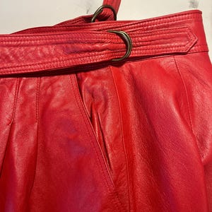 May include: Close-up of a red leather skirt with a belt. The skirt has a high waist and a belt with a metal buckle. The leather appears to be soft and supple. The skirt has a classic, vintage style.