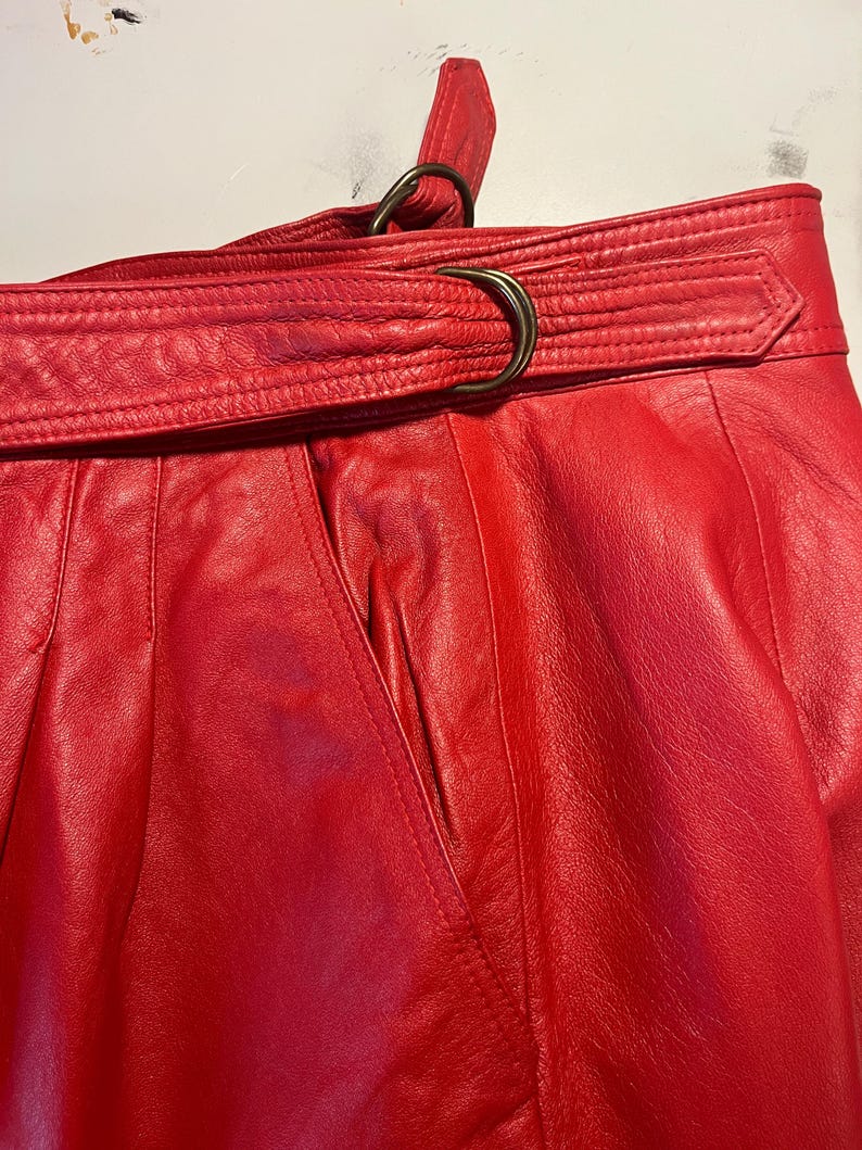 May include: Close-up of a red leather skirt with a belt. The skirt has a high waist and a belt with a metal buckle. The leather appears to be soft and supple. The skirt has a classic, vintage style.