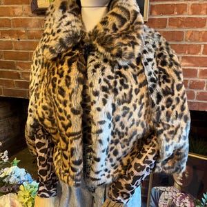 K&ouml;nnte beinhalten: Ein kurzes Kunstfell-Cape mit einem auff&auml;lligen Leopardenmuster in Beige-, Braun- und Schwarzt&ouml;nen. Das Kleidungsst&uuml;ck hat einen breiten Kragen, einen einzelnen Knebelverschluss am Ausschnitt und wird &uuml;ber einem lang&auml;rmeligen Leoparden-Oberteil und einer hellblauen Jeans getragen.