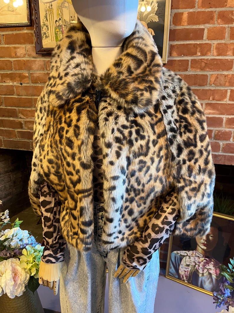 K&ouml;nnte beinhalten: Ein kurzes Kunstfell-Cape mit einem auff&auml;lligen Leopardenmuster in Beige-, Braun- und Schwarzt&ouml;nen. Das Kleidungsst&uuml;ck hat einen breiten Kragen, einen einzelnen Knebelverschluss am Ausschnitt und wird &uuml;ber einem lang&auml;rmeligen Leoparden-Oberteil und einer hellblauen Jeans getragen.