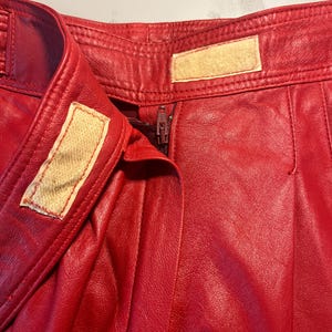 May include: Close-up of red leather pants. The pants have a zipper closure and a button closure. The leather appears to be slightly textured. The pants have a high waist and a classic design.