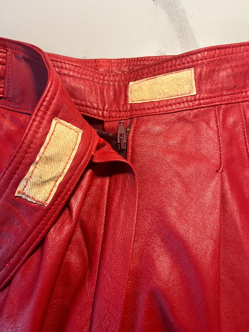 May include: Close-up of red leather pants. The pants have a zipper closure and a button closure. The leather appears to be slightly textured. The pants have a high waist and a classic design.