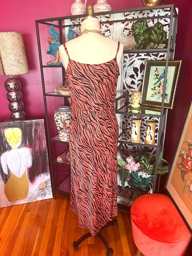 Puede incluir: Vestido lencero color coral con estampado de cebra negro. El vestido tiene tirantes finos y largo midi. Se exhibe en un maniqu&iacute; frente a una estanter&iacute;a de metal negro con objetos decorativos.