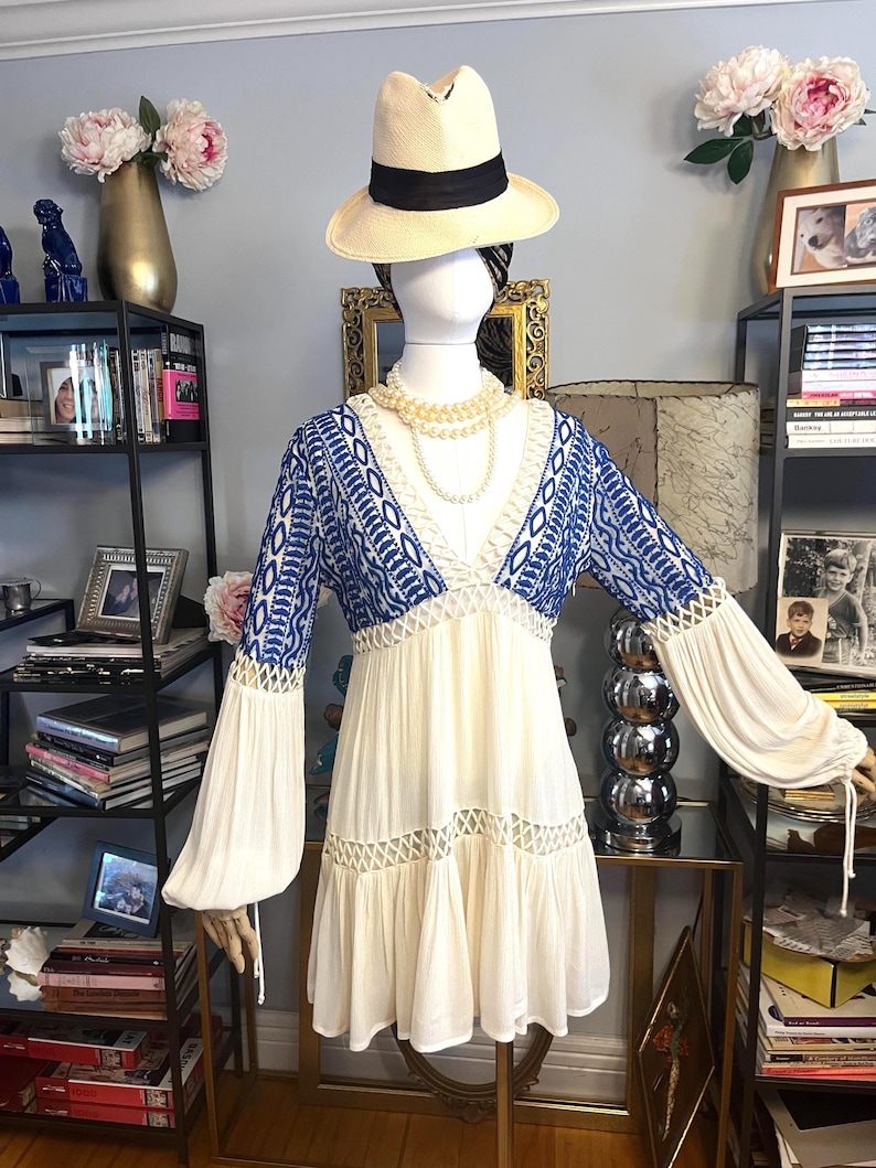 Pu&ograve; includere: Un abito color crema con dettagli ricamati blu sul corpetto e sulle maniche. L'abito presenta uno scollo a V, maniche lunghe e fluide e una gonna a balze. Un cappello di paglia con una fascia nera e una collana di perle completano il look. L'abito &egrave; esposto su un manichino.