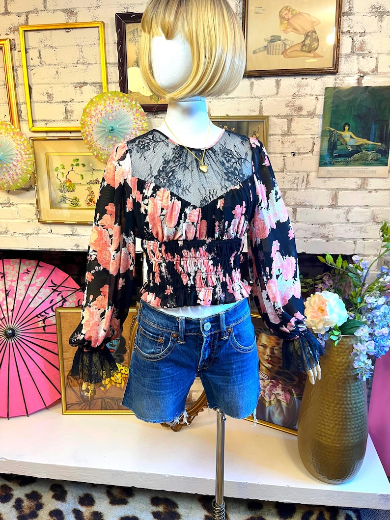 May include: A black floral print blouse with pink flowers and a lace neckline is paired with denim shorts. The blouse has long sleeves with black lace cuffs. A gold necklace with a heart pendant is visible.