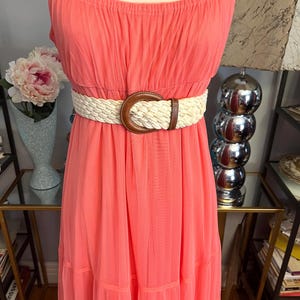 Puede incluir: Un vestido largo color coral con tirantes finos y un cintur&oacute;n trenzado. El cintur&oacute;n tiene una hebilla de cuero marr&oacute;n. El vestido est&aacute; hecho de una tela ligera y tiene una falda escalonada. Un collar de perlas es visible en la parte superior.