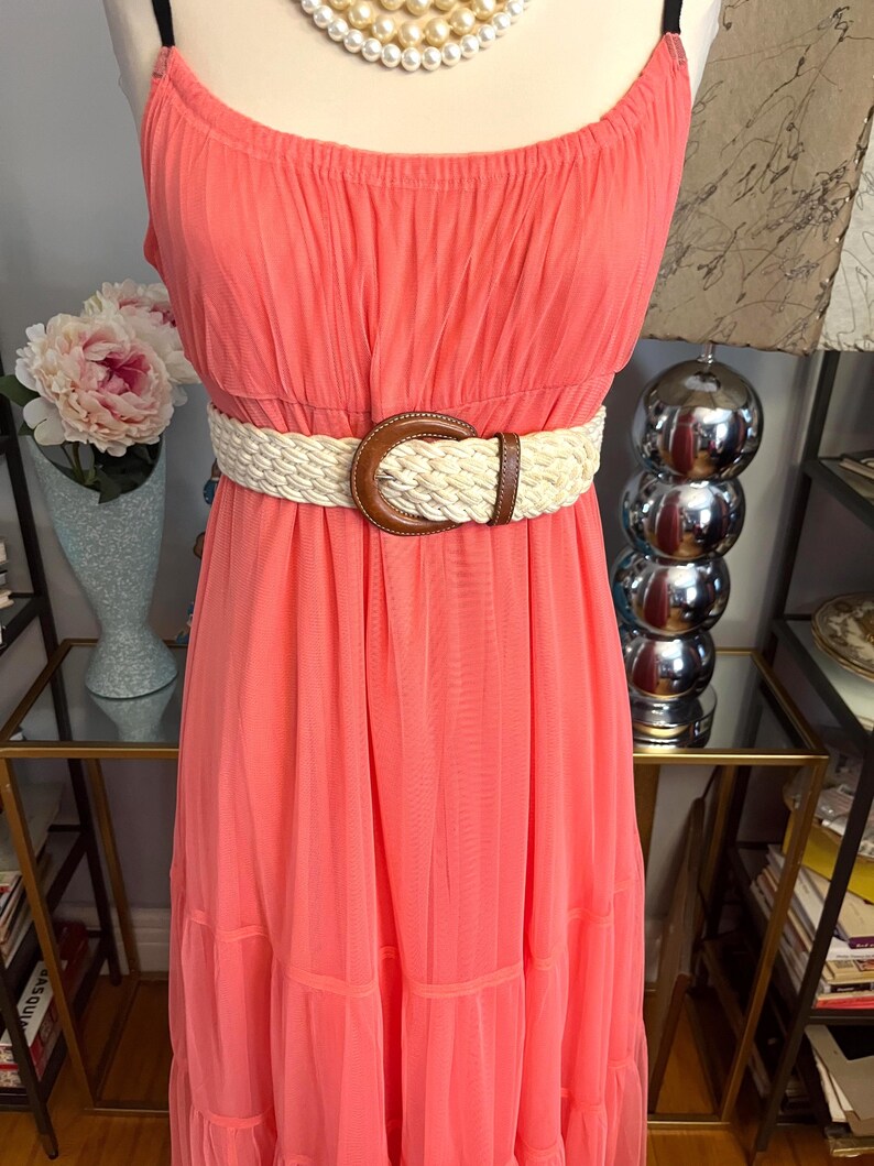 Puede incluir: Un vestido largo color coral con tirantes finos y un cintur&oacute;n trenzado. El cintur&oacute;n tiene una hebilla de cuero marr&oacute;n. El vestido est&aacute; hecho de una tela ligera y tiene una falda escalonada. Un collar de perlas es visible en la parte superior.