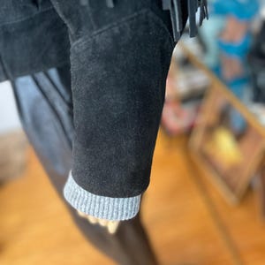 May include: Close-up of a black suede jacket with fringe detailing. The jacket features a gray ribbed knit cuff on the sleeve. The garment is displayed on a mannequin.