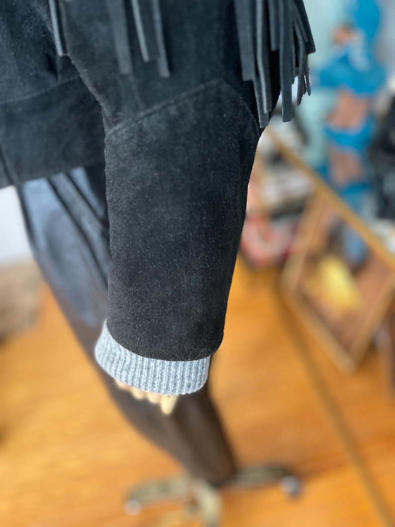 May include: Close-up of a black suede jacket with fringe detailing. The jacket features a gray ribbed knit cuff on the sleeve. The garment is displayed on a mannequin.