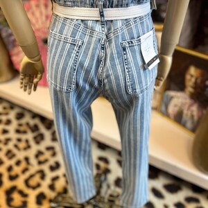 Pode incluir: Jeans de ganga azul claro com riscas brancas verticais. Os jeans t&ecirc;m cintura alta, cinto branco e bolsos traseiros. Os jeans s&atilde;o exibidos num manequim.