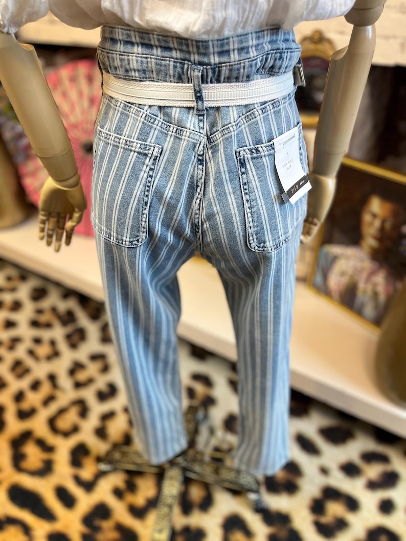 Pode incluir: Jeans de ganga azul claro com riscas brancas verticais. Os jeans t&ecirc;m cintura alta, cinto branco e bolsos traseiros. Os jeans s&atilde;o exibidos num manequim.