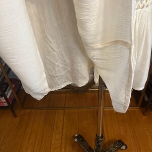 Pu&ograve; includere: Un capo di abbigliamento in tessuto bianco sporco, strutturato, possibilmente un vestito o un top, appeso a un supporto metallico decorativo. Il tessuto ha un aspetto stropicciato e una lucentezza sottile. Il capo presenta un dettaglio all'uncinetto.