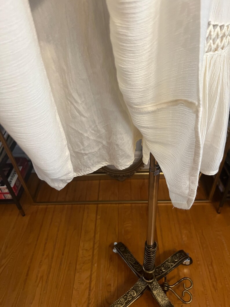 Pu&ograve; includere: Un capo di abbigliamento in tessuto bianco sporco, strutturato, possibilmente un vestito o un top, appeso a un supporto metallico decorativo. Il tessuto ha un aspetto stropicciato e una lucentezza sottile. Il capo presenta un dettaglio all'uncinetto.