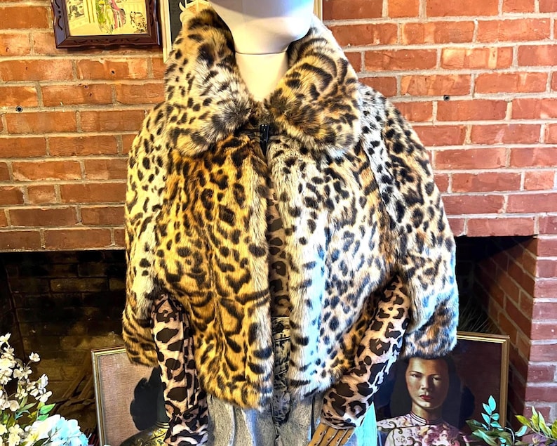 K&ouml;nnte beinhalten: Eine kurz geschnittene Jacke mit einem auff&auml;lligen Leopardenmuster in Braun-, Schwarz- und Beiget&ouml;nen. Das Kleidungsst&uuml;ck hat einen breiten, flauschigen Kragen und lange &Auml;rmel mit einer fell&auml;hnlichen Textur und wird &uuml;ber einem passenden Oberteil getragen.