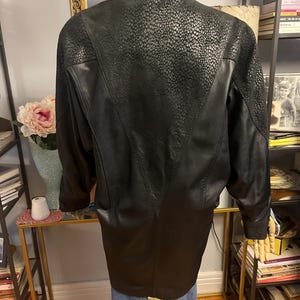 May include: Black leather jacket with a high collar and textured detailing on the upper back. The jacket has a long, tailored fit and features a V-shaped design on the back. The sleeves are long and the jacket appears to be vintage.