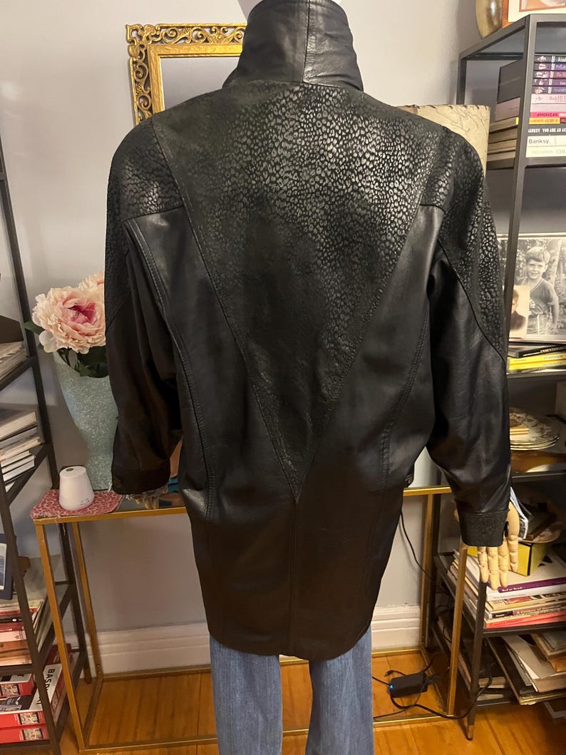 May include: Black leather jacket with a high collar and textured detailing on the upper back. The jacket has a long, tailored fit and features a V-shaped design on the back. The sleeves are long and the jacket appears to be vintage.