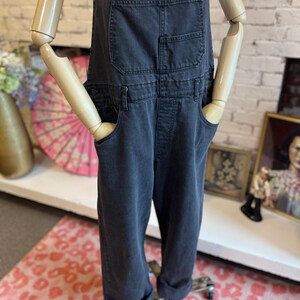 May include: Dark gray denim overalls with front and back pockets. The overalls have adjustable straps and a relaxed fit. The cuffs are rolled up. The overalls are displayed on a mannequin.
