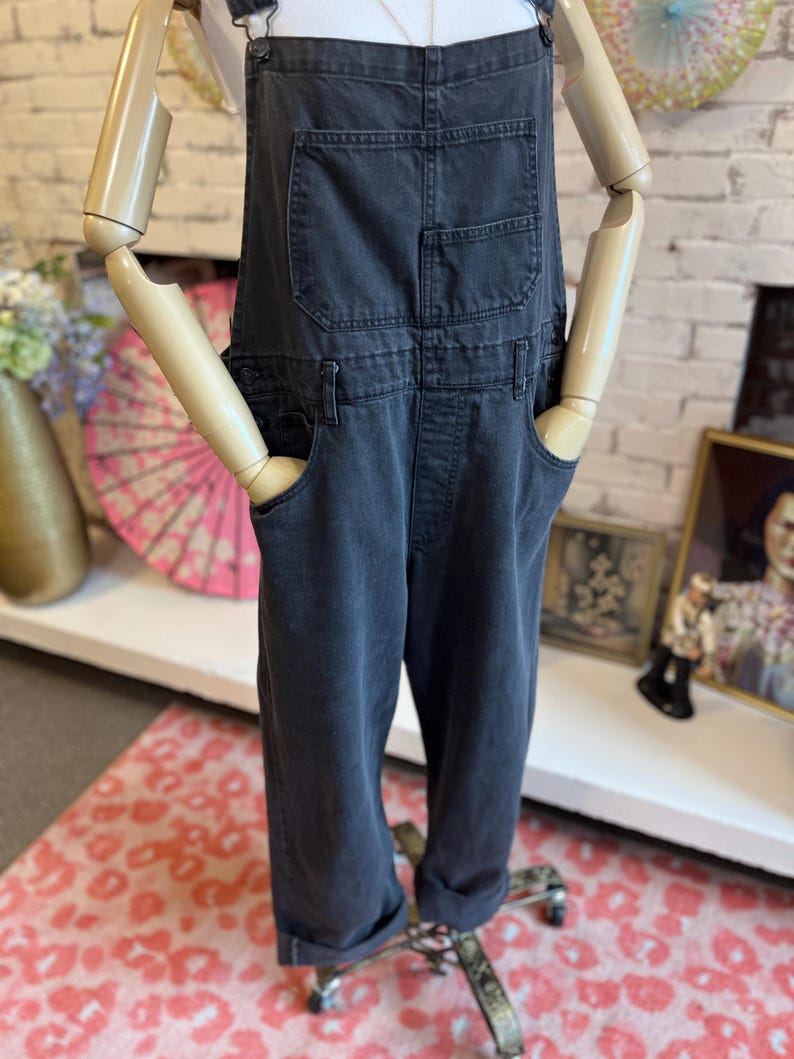 May include: Dark gray denim overalls with front and back pockets. The overalls have adjustable straps and a relaxed fit. The cuffs are rolled up. The overalls are displayed on a mannequin.