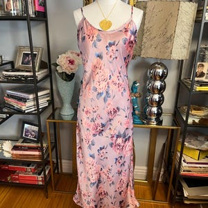 May include: A light purple slip dress with a floral pattern of pink and blue flowers. The dress has thin straps and a flowing silhouette. A gold pendant necklace hangs on the mannequin. The dress is displayed on a mannequin.