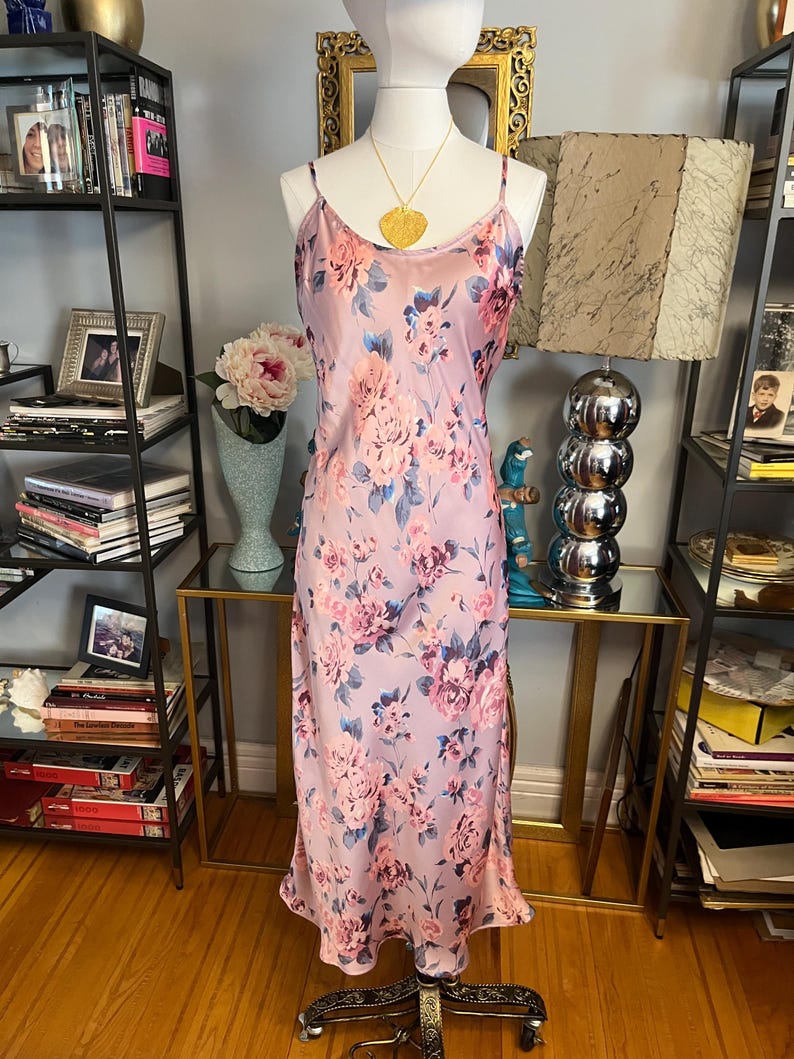 May include: A light purple slip dress with a floral pattern of pink and blue flowers. The dress has thin straps and a flowing silhouette. A gold pendant necklace hangs on the mannequin. The dress is displayed on a mannequin.