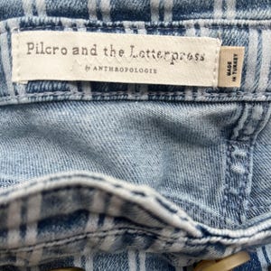 Pode incluir: Close-up de tecido denim azul claro com um padr&atilde;o xadrez branco. A etiqueta diz "Pilcro and the Letterpress by Anthropologie" e "Made in Turkey". Uma fivela de cinto dourada e um bot&atilde;o s&atilde;o vis&iacute;veis.