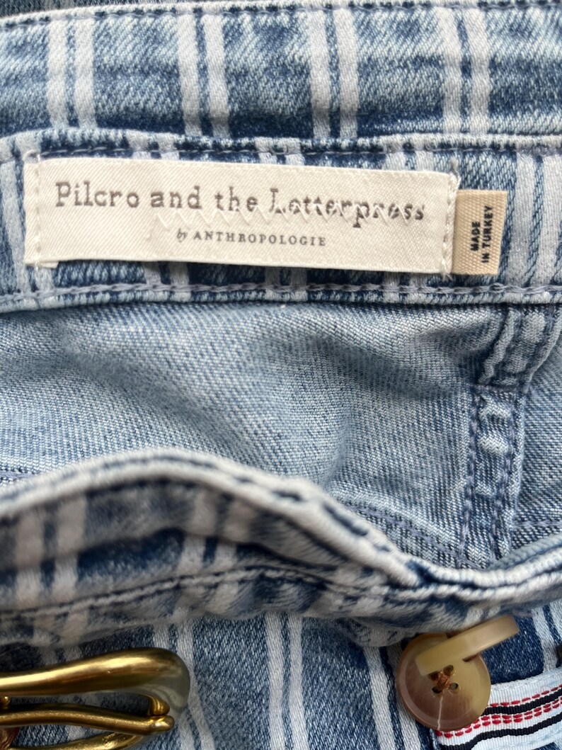 Pode incluir: Close-up de tecido denim azul claro com um padr&atilde;o xadrez branco. A etiqueta diz "Pilcro and the Letterpress by Anthropologie" e "Made in Turkey". Uma fivela de cinto dourada e um bot&atilde;o s&atilde;o vis&iacute;veis.