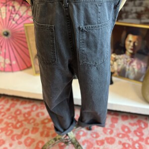 May include: Dark gray denim overalls with two back pockets and rolled-up cuffs. The overalls are displayed on a mannequin. The fabric appears to be a medium weight denim. The overall style is casual and relaxed.