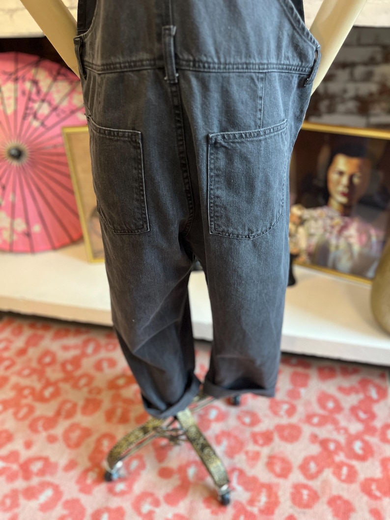 May include: Dark gray denim overalls with two back pockets and rolled-up cuffs. The overalls are displayed on a mannequin. The fabric appears to be a medium weight denim. The overall style is casual and relaxed.