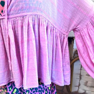May include: A pink blouse with a ruffled hem and long, flared sleeves. The top has a textured, woven appearance and a drawstring neckline. The lower portion of the image shows a skirt with a leopard print design in pink, green, and black.